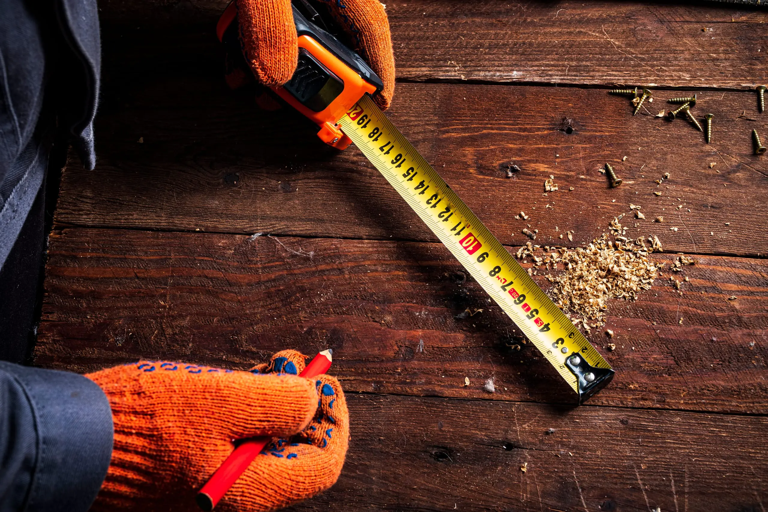 Gloved hands measuring a wooden board with a tape measure and a carpenter’s pencil